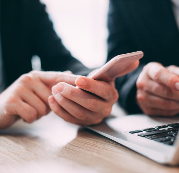 Close-up of hand of businesswoman holding mobile phone and reading message. Male colleague sitting nearby and using laptop